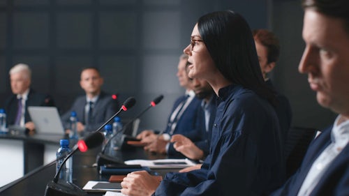 Eine Frau sitzt und spricht vor einem Regulierungsgremium, umgeben von Kollegen